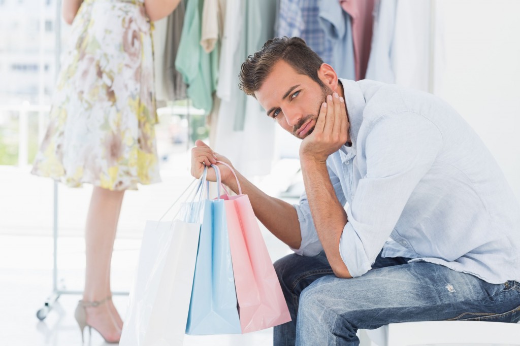 man with shopping bag bored while woman searches clothes rack