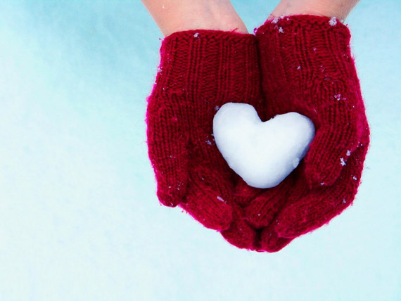 pair of cupped hands in red mittens holding heart-shaped snowball against a white and blue background